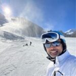 Person in white ski gear and goggles taking a selfie on a sunny snow-covered mountain slope, with skiers in the background.